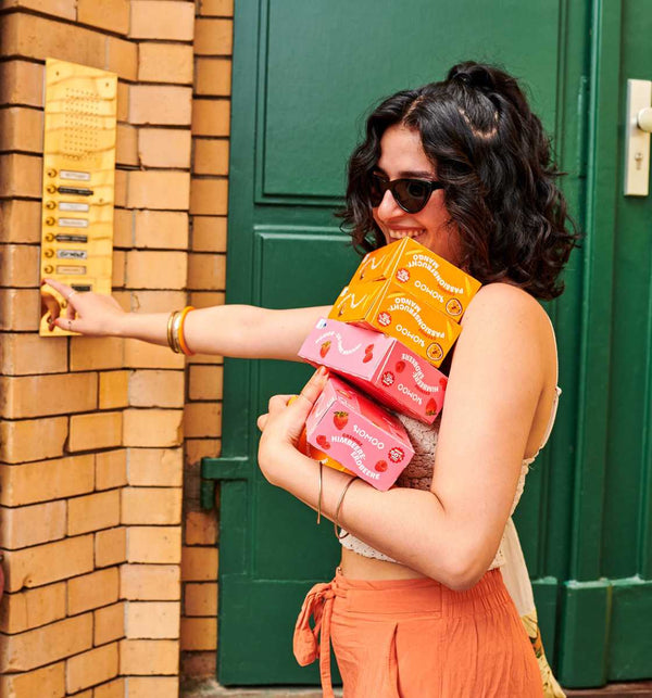 Frau klingelt an Haustür mit Mango-Passionsfrucht und Erdbeere-Himbeere Flowpacks im Arm